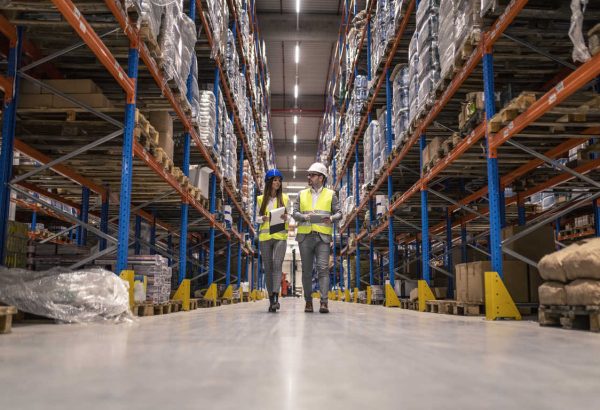 Workers with hardhats and reflective jackets walking through big warehouse aisle checking goods condition. Workers with hardhats and reflective jackets walking through big warehouse aisle checking goods condition.